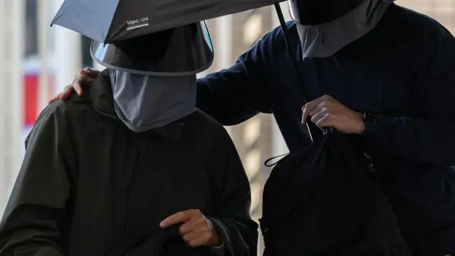 两名身穿黑色外套、戴黑色帽子及口罩的男子共撑一把雨伞。